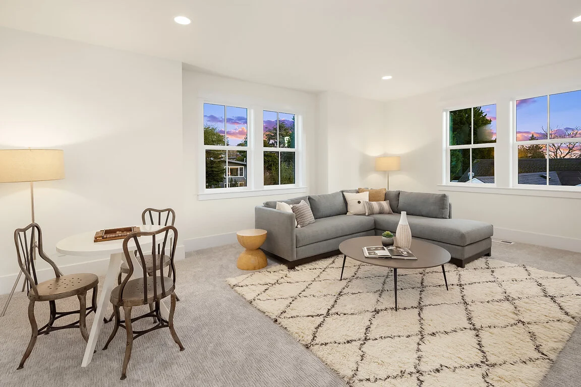 Living room with a gray sectional sofa, white walls, multiple windows showing an evening sky, a round coffee table with decorative vases, and a white area rug with a diamond pattern.