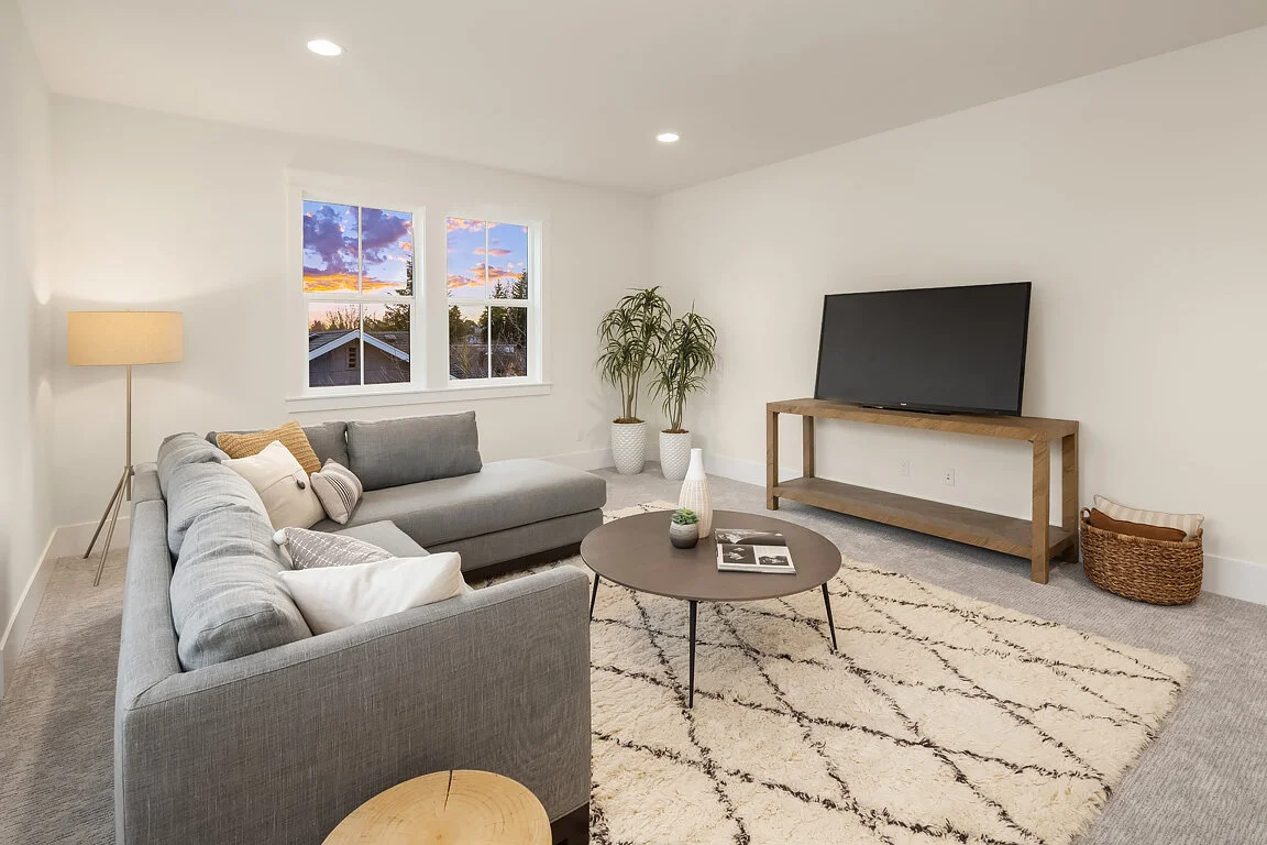 Living room with gray sectional sofa, round coffee table, potted plants, large window showing a sunset sky, and a flat-screen TV on a wooden stand.
