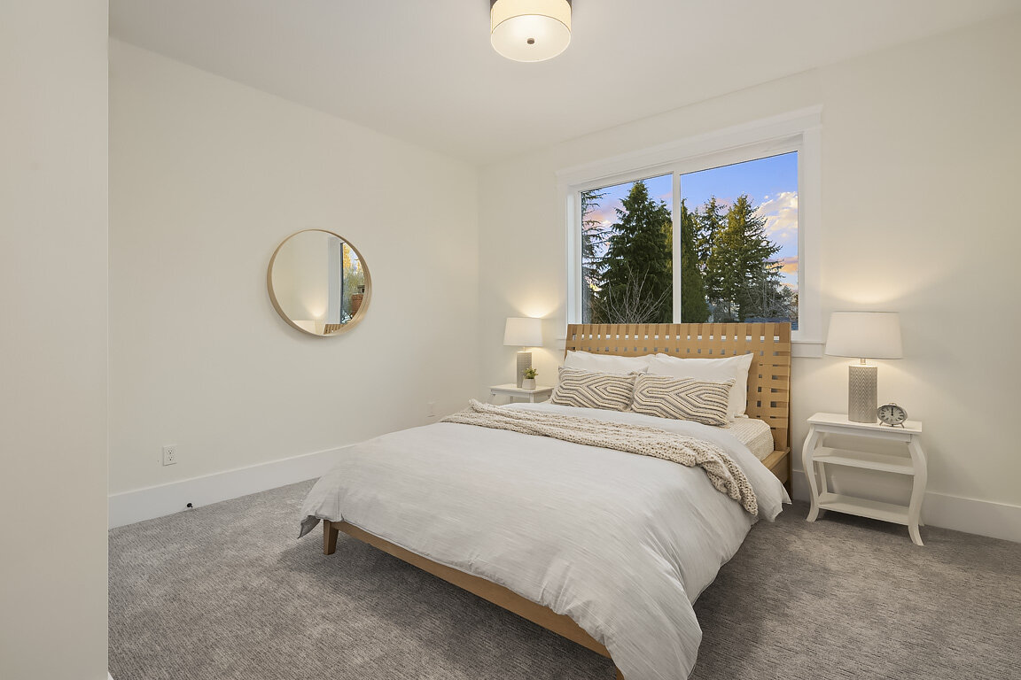 A minimalist bedroom with a bed, two bedside tables, table lamps, a round mirror on the wall, and a window showing trees outside at sunset.