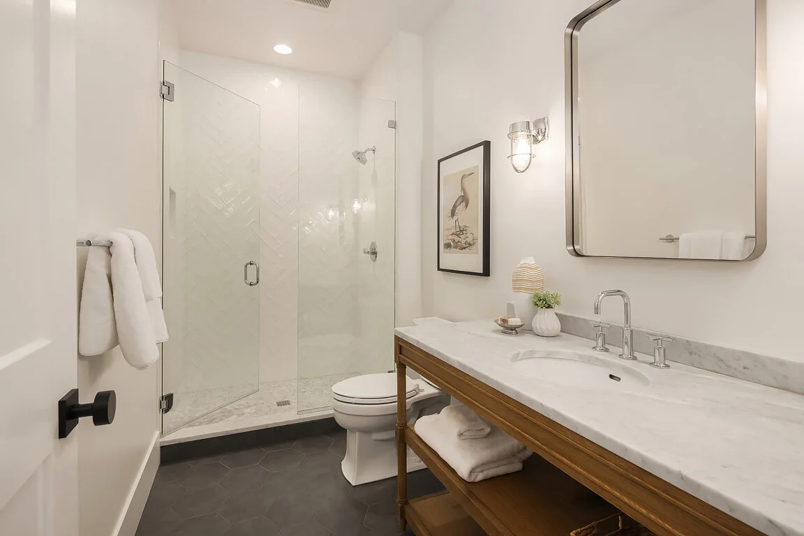Modern bathroom with walk-in glass shower, white toilet, marble countertop with sink, wooden vanity, framed art, towel rack with towels, and decorative items.