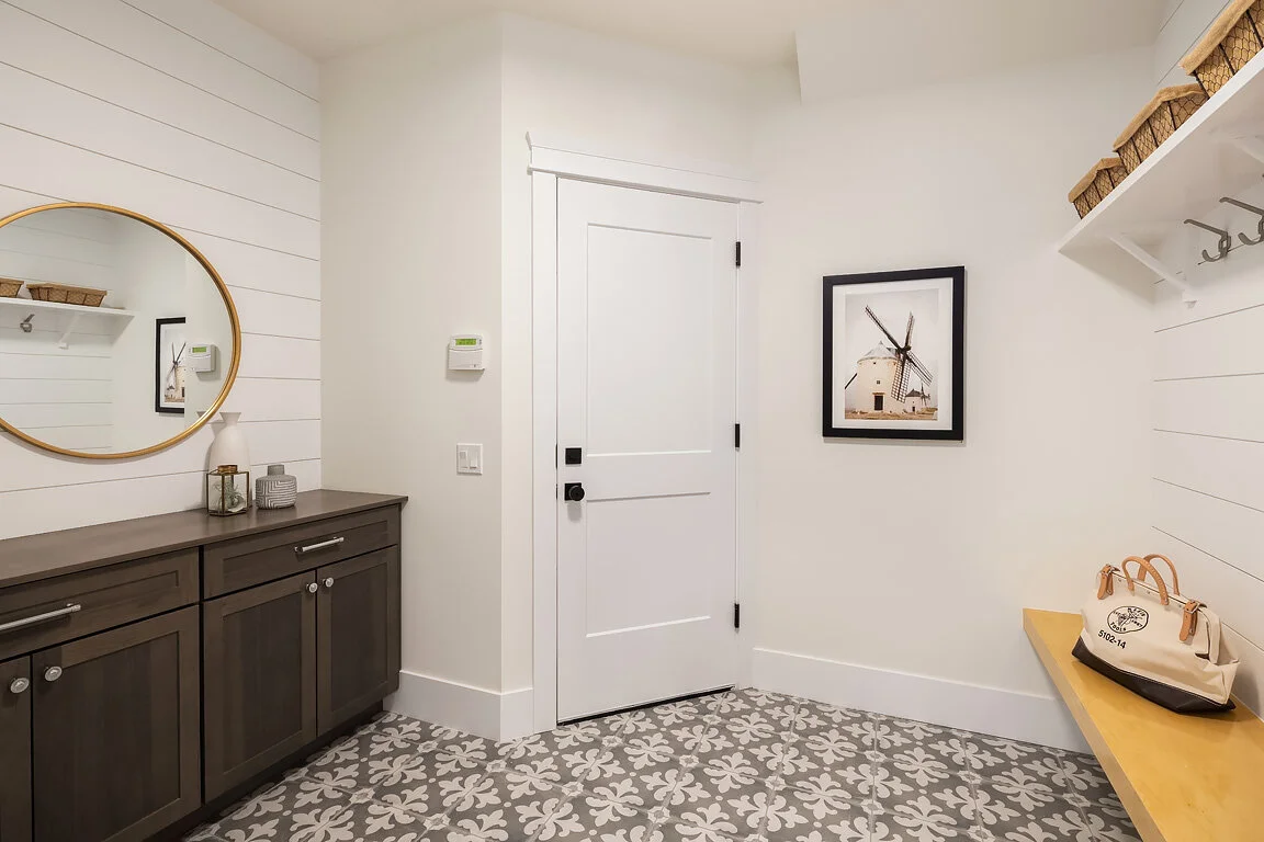 Entryway with patterned tile floor, white paneled walls, a white door, a gray sideboard with a mirror, decorative items, framed art on the wall, baskets on a white shelf, and a beige bag on a yellow bench.