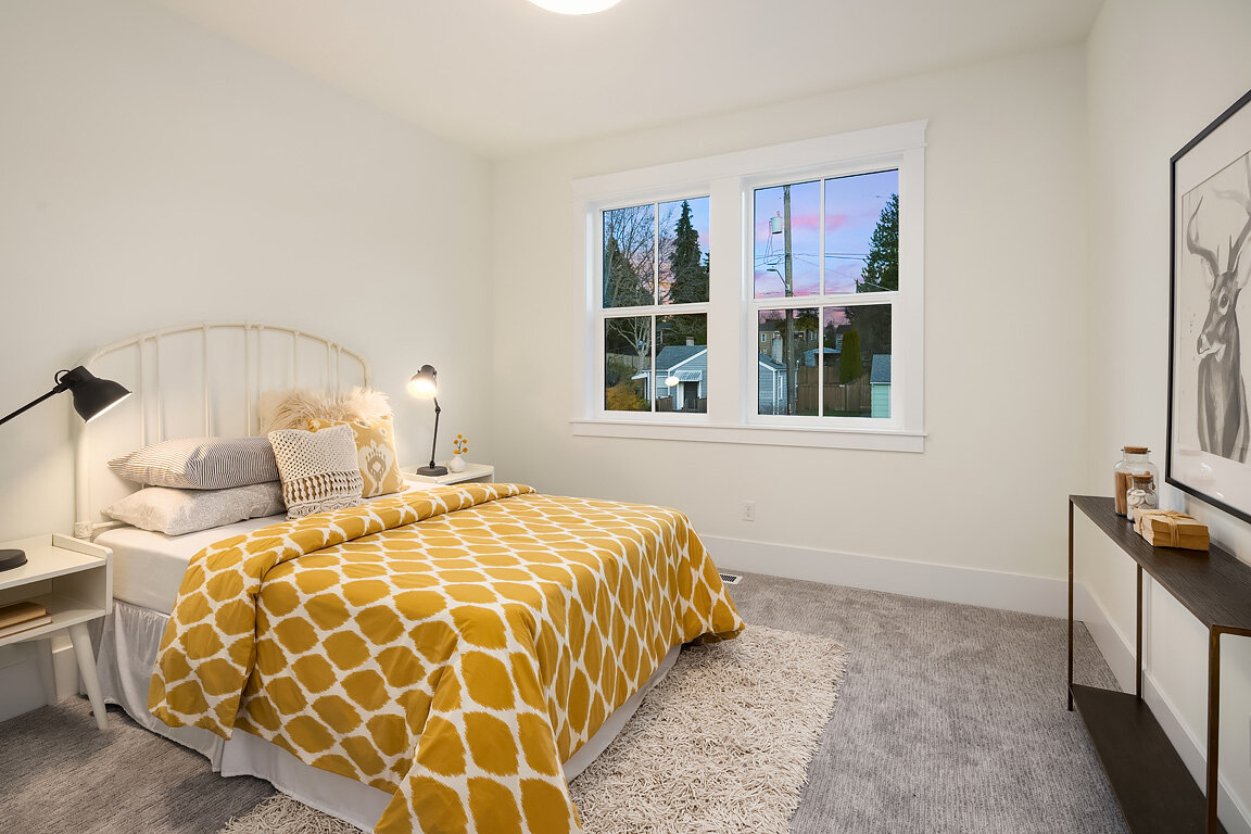 A bedroom with white walls, a window showing trees and houses outside, a bed with a yellow patterned duvet, and black lamps on white bedside tables.