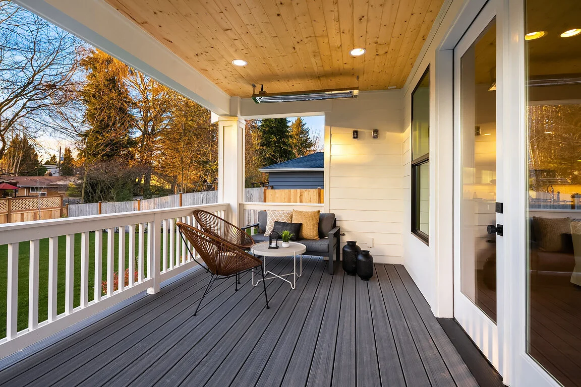 A spacious outdoor balcony with wooden flooring and a wooden ceiling. It has a seating area with a black sofa, two brown wicker chairs, and a small white round table with decorative items. The balcony overlooks a grassy yard with trees, surrounded by