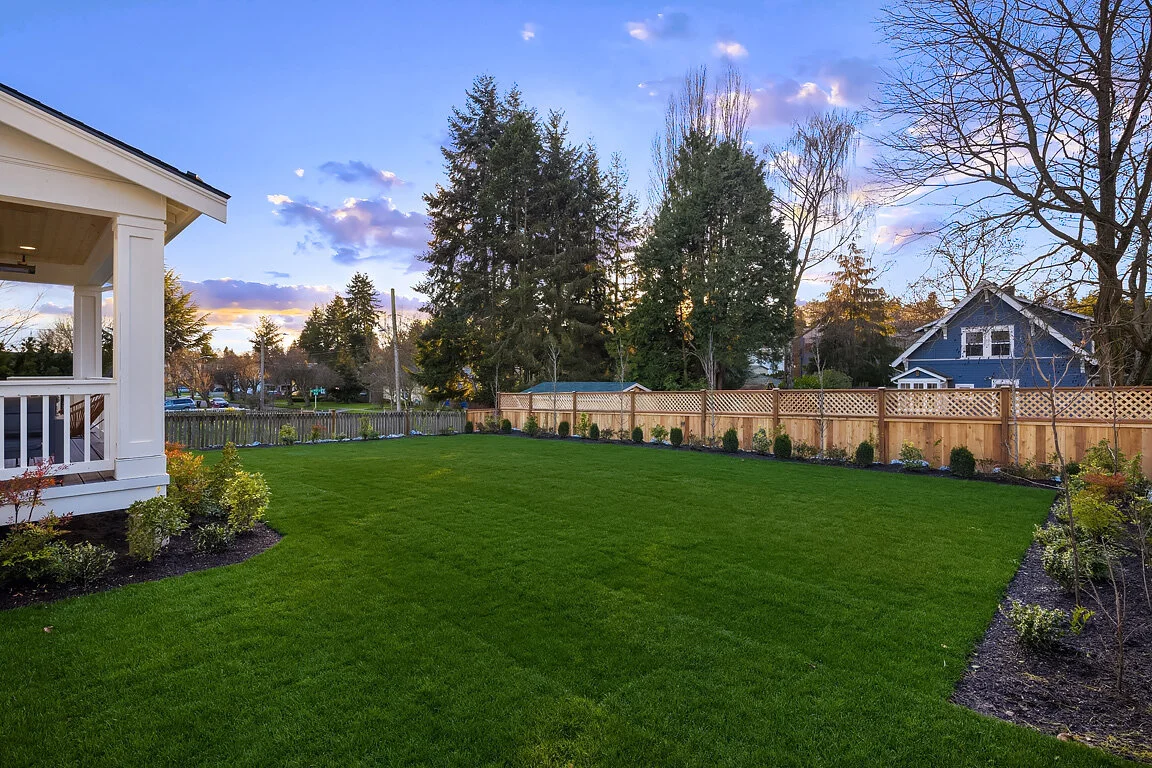 A backyard with a well-maintained green lawn, a wooden fence, and a house on the left with a porch. The sky is partly cloudy during sunset or sunrise, and there are trees and neighboring houses in the background.