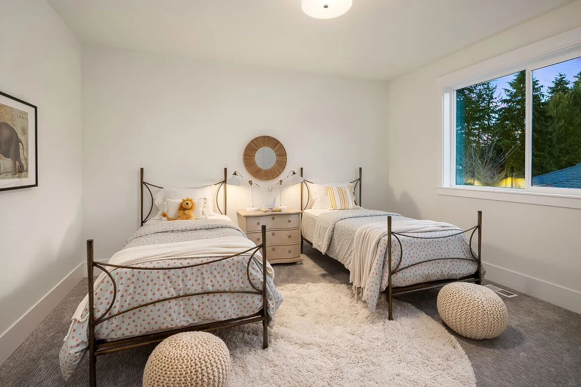 A bedroom with two twin beds, a white nightstand in between, a mirror above the nightstand, and a large window showing trees outside. The beds havebed linens with a small pattern and pillows, one with a teddy bear. There are two knitted poufs on the 