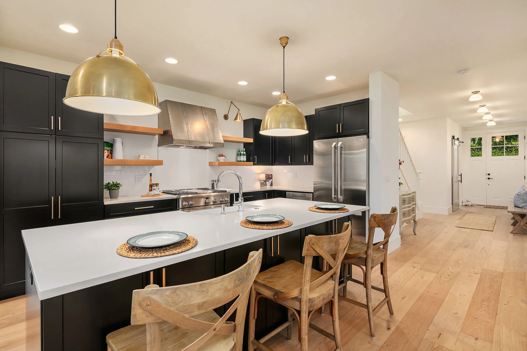 Modern kitchen with black cabinets, white countertops, gold pendant lights, open wooden shelves, stainless steel appliances, and wooden chairs at the island.