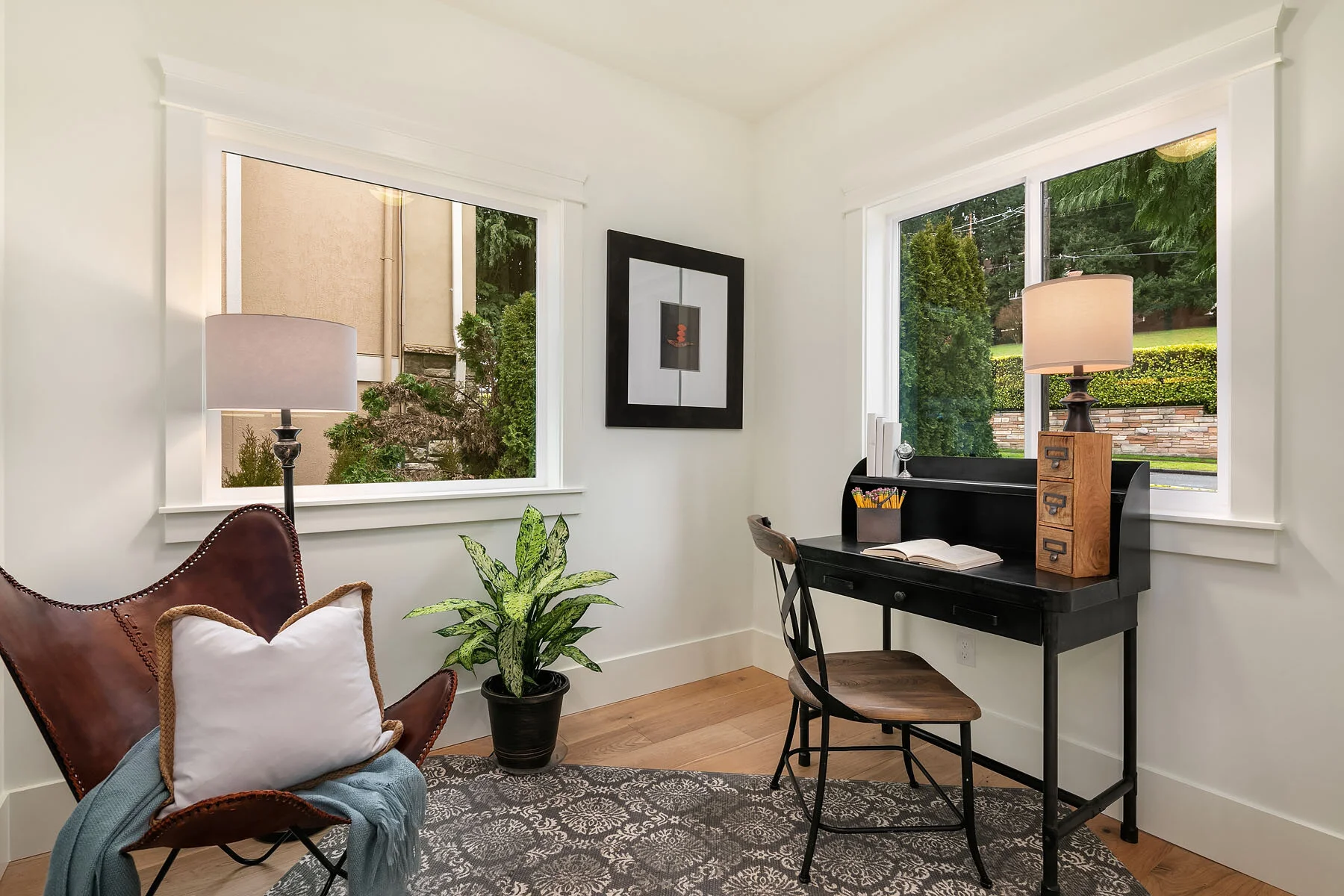 A cozy home office with a black desk, wooden chair, and a desk lamp. There is a brown butterfly chair with a white pillow, a green potted plant, and two windows overlooking a green yard.
