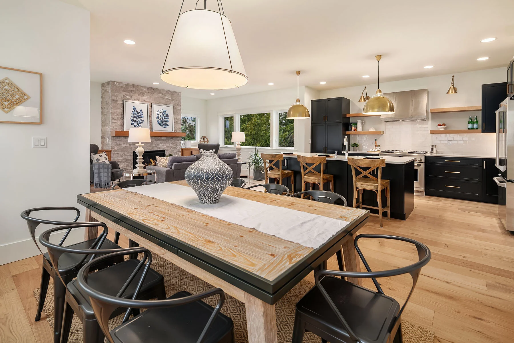 Open-concept kitchen and dining area with a wooden table, black chairs, and a decorative vase in the foreground. The kitchen features black cabinets, a white subway tile backsplash, a breakfast bar with wooden stools, and brass pendant lights. The ba