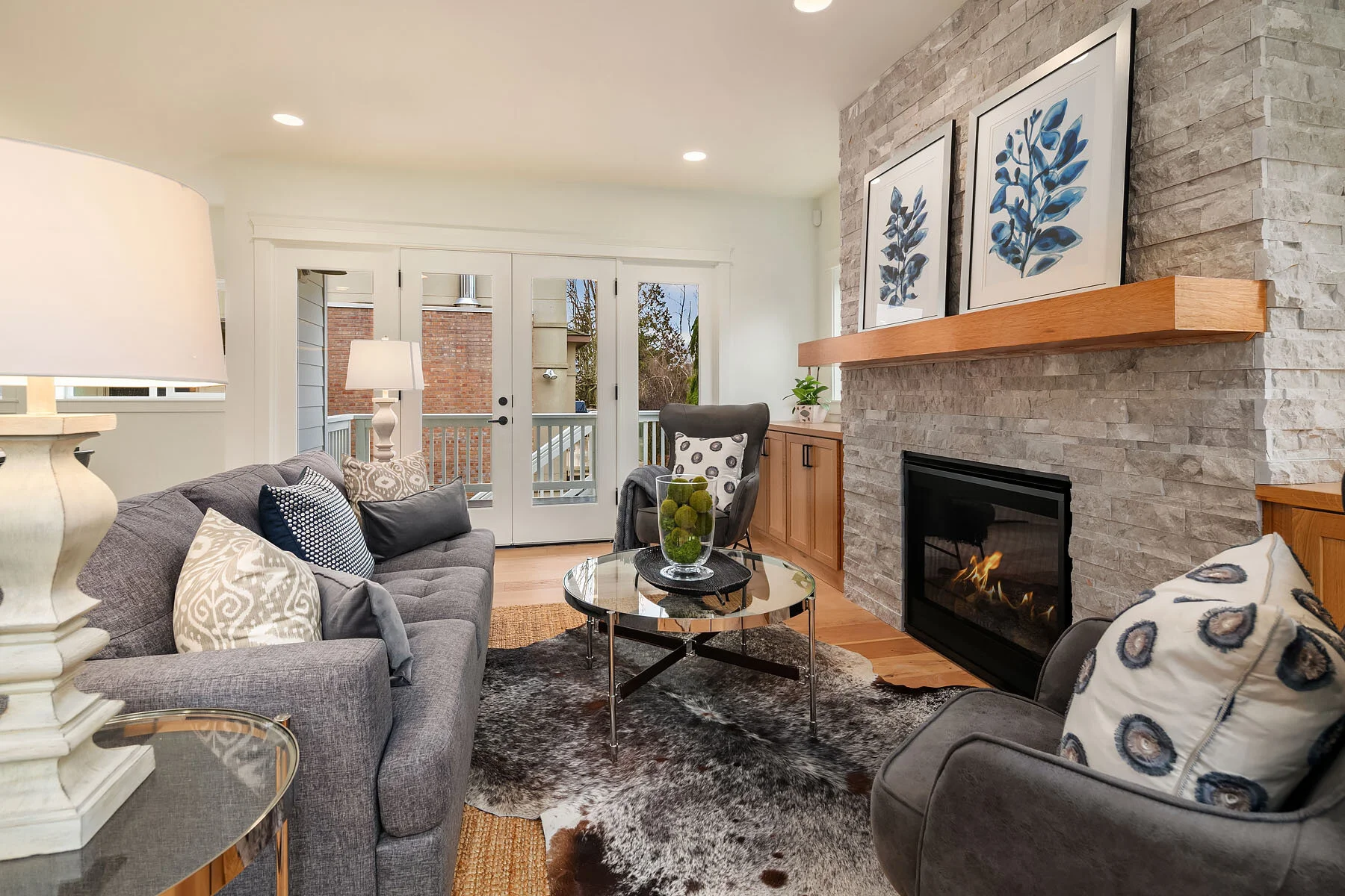 Living room with grey sofa, patterned pillows, glass coffee table, sliding glass door leading outside, mounted artwork above fireplace, armchair with decorative pillow, and wood accents.