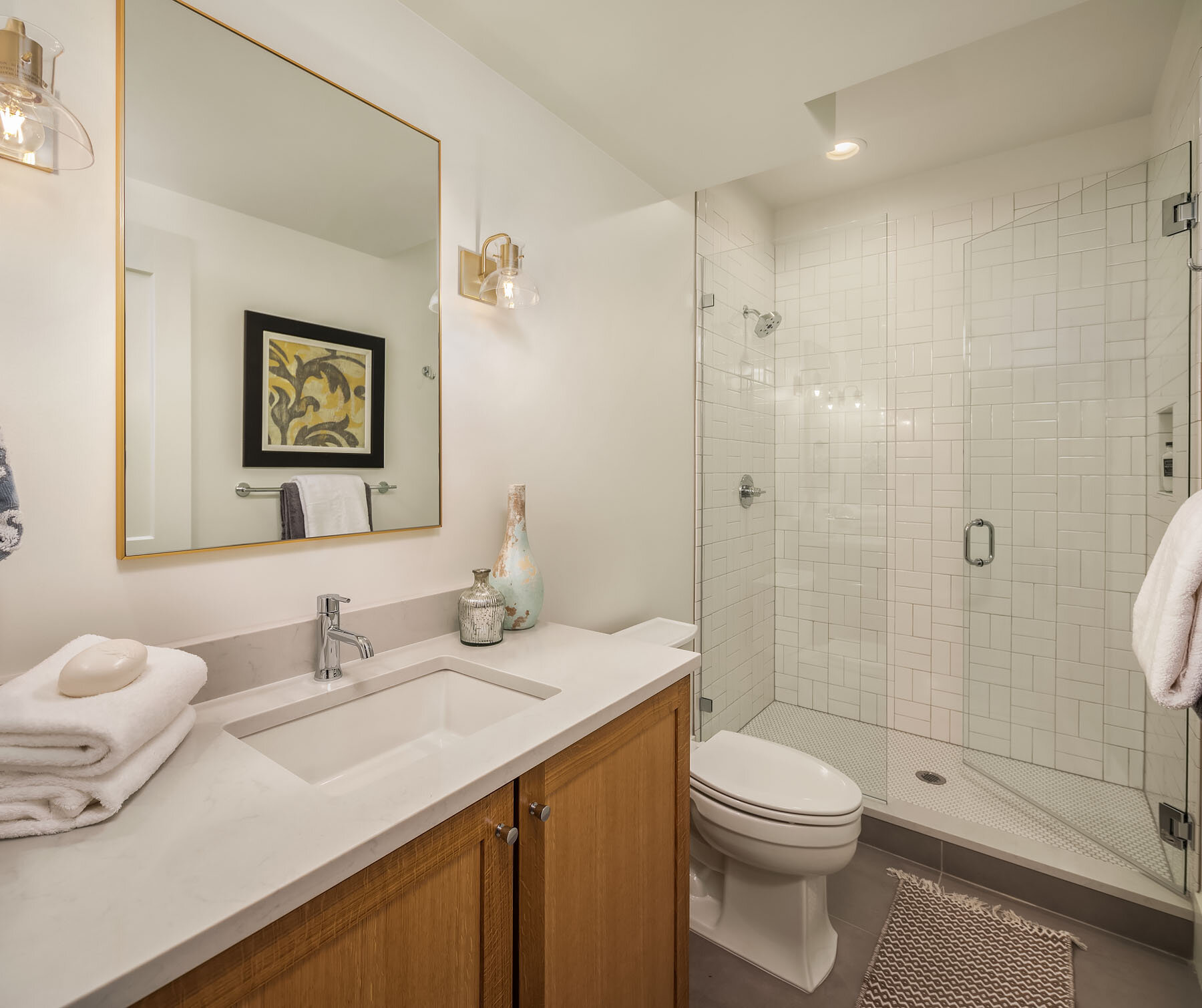 Modern bathroom with a white vanity, a mirror, towels, decorative vases, a toilet, and a glass-enclosed shower with white subway tiles.