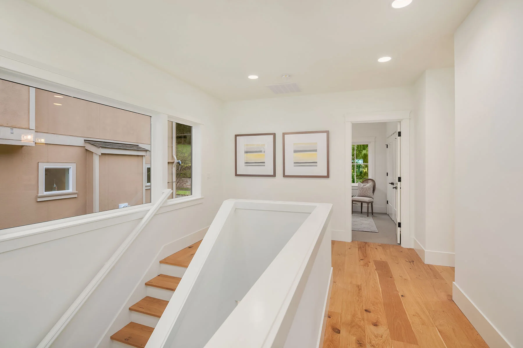Interior view of a modern home with white walls, hardwood floors, a staircase, large windows, and framed artwork on the wall.