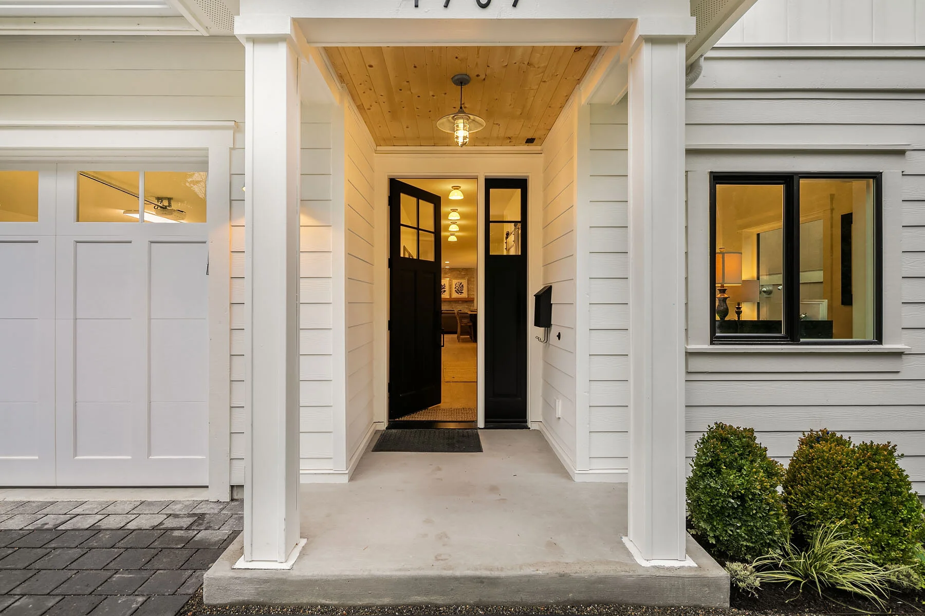 The front porch of a modern house with white siding, black front door, and a window showing a lit lamp inside. Small shrubs are planted near the porch.