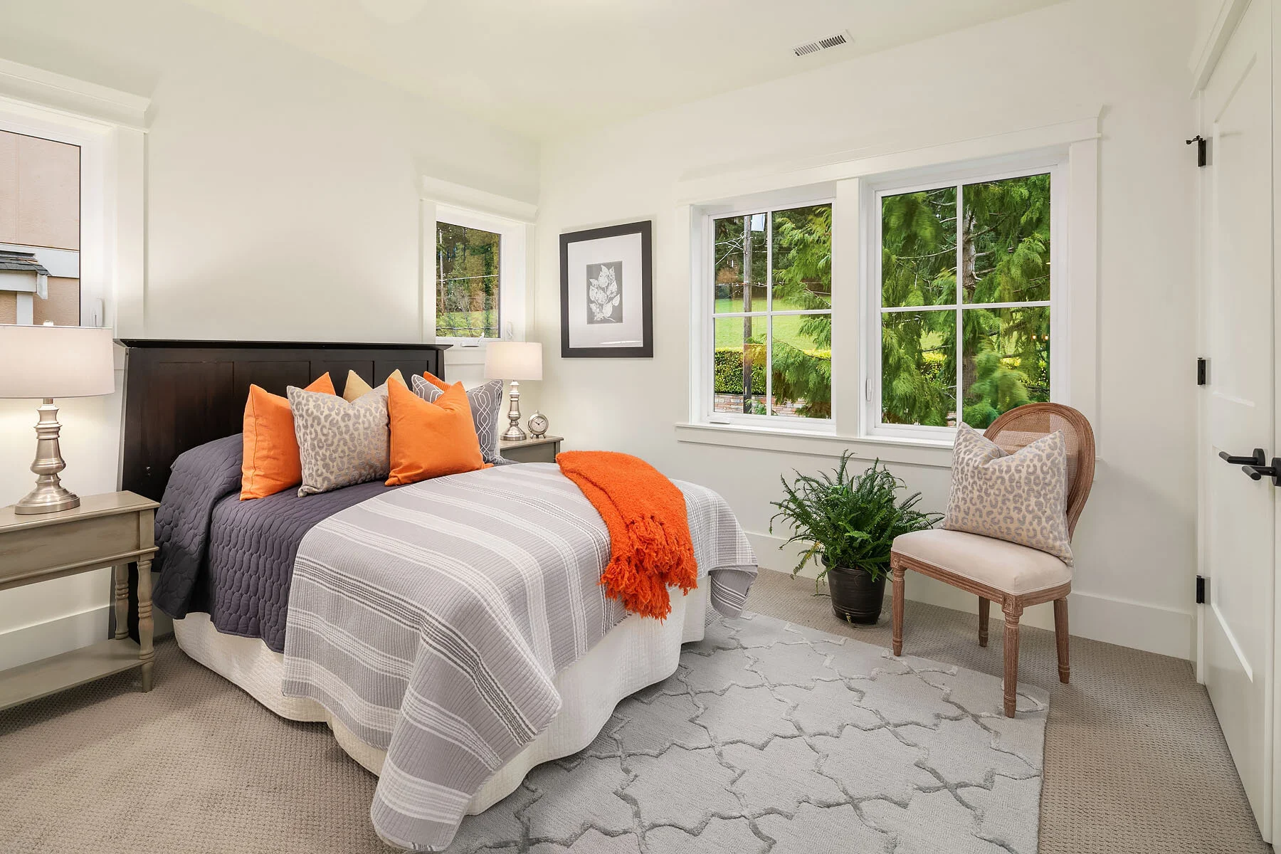 A cozy bedroom with a large bed, multiple colorful pillows, and a throw blanket. There are two nightstands with lamps, a framed black and white artwork, a window showing greenery outside, and a chair with a pillow near a potted plant.