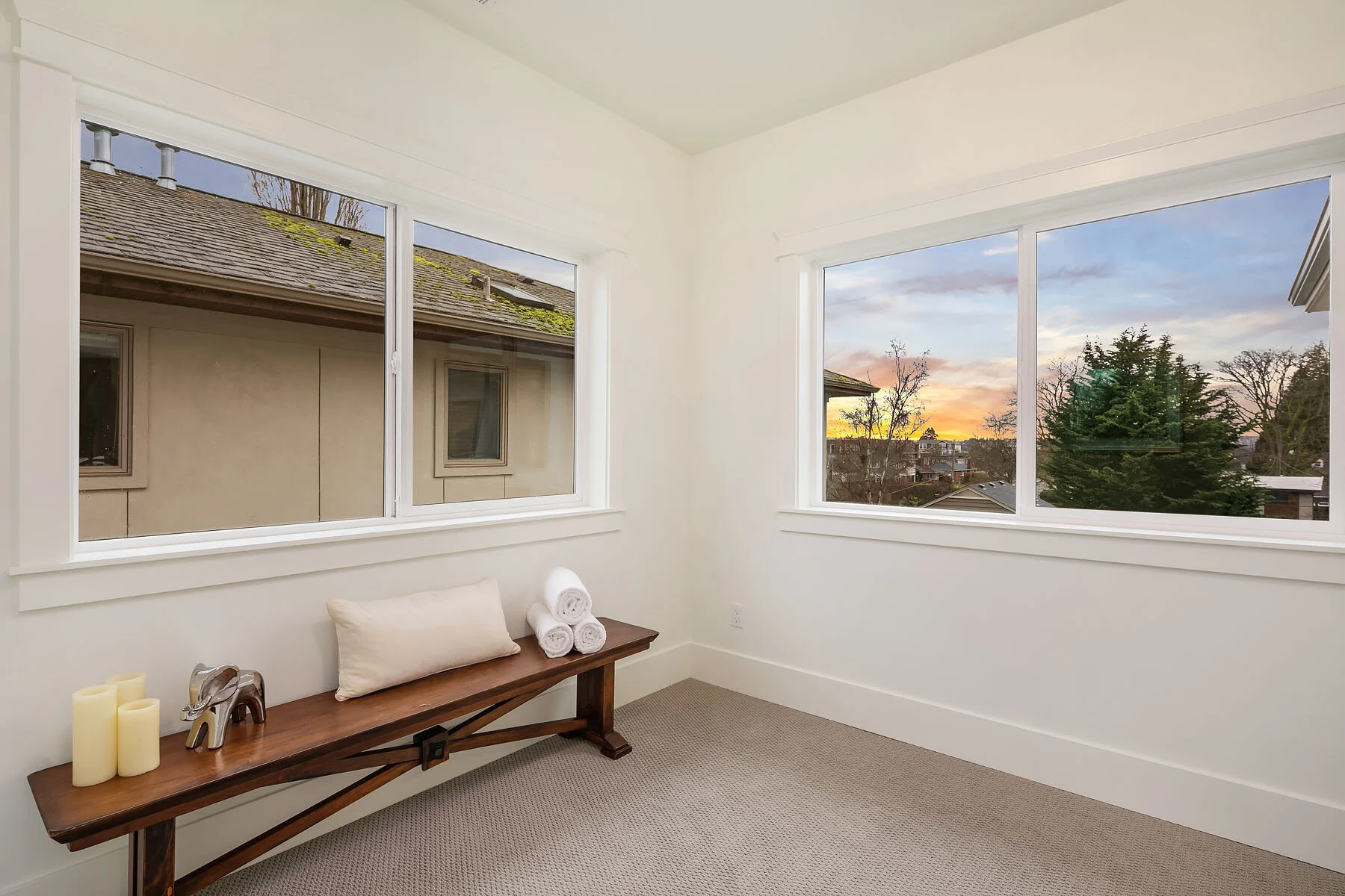 Empty room with large windows showing a sunset sky and trees outside, a wooden bench with candles, a pillow, and rolled towels inside.