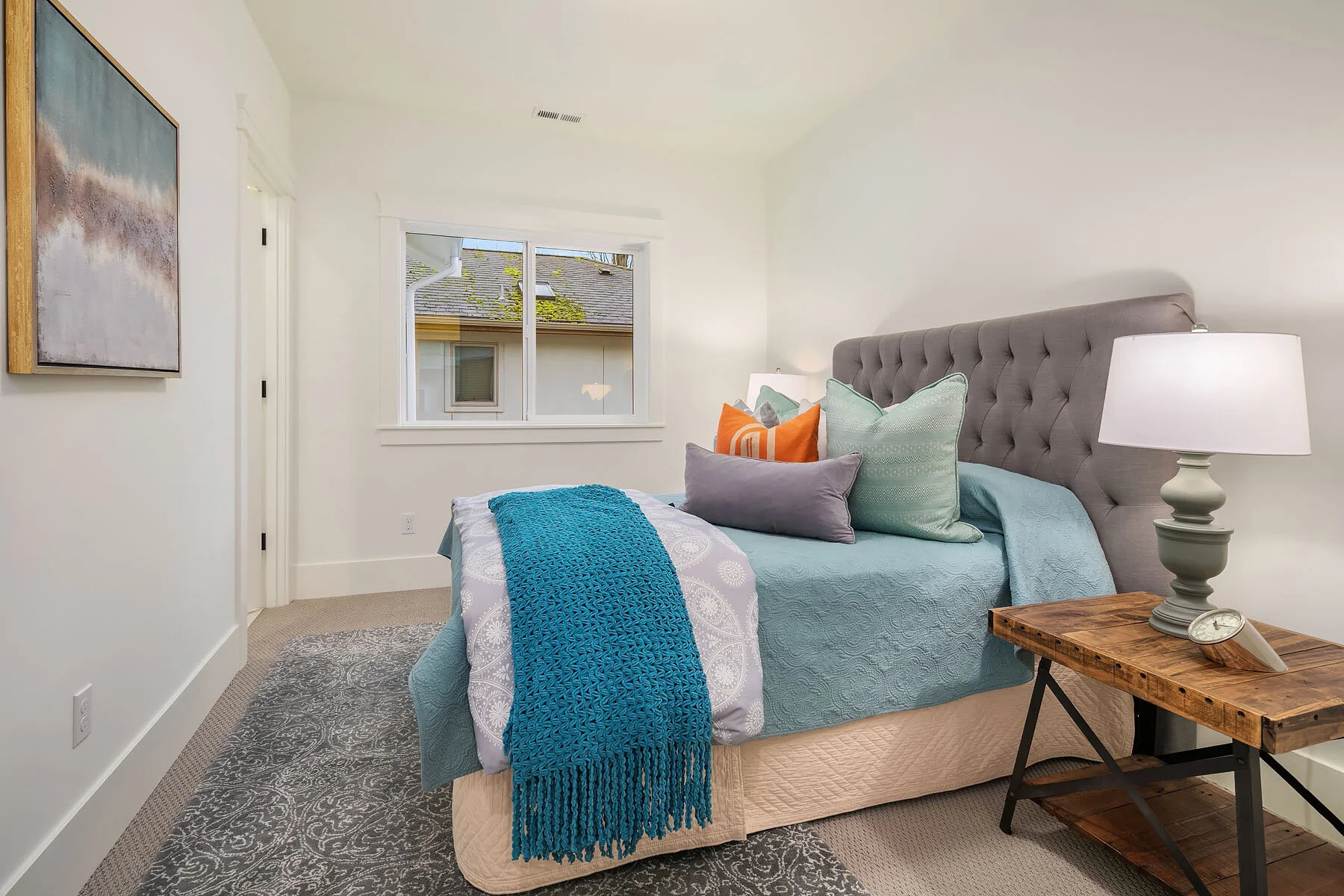 A bedroom featuring a bed with a tufted gray headboard, multiple pillows, a blue bedspread, and a knitted blue throw. A wooden nightstand with a lamp and clock is next to the bed. There is a window in the background with a view of neighboring houses 