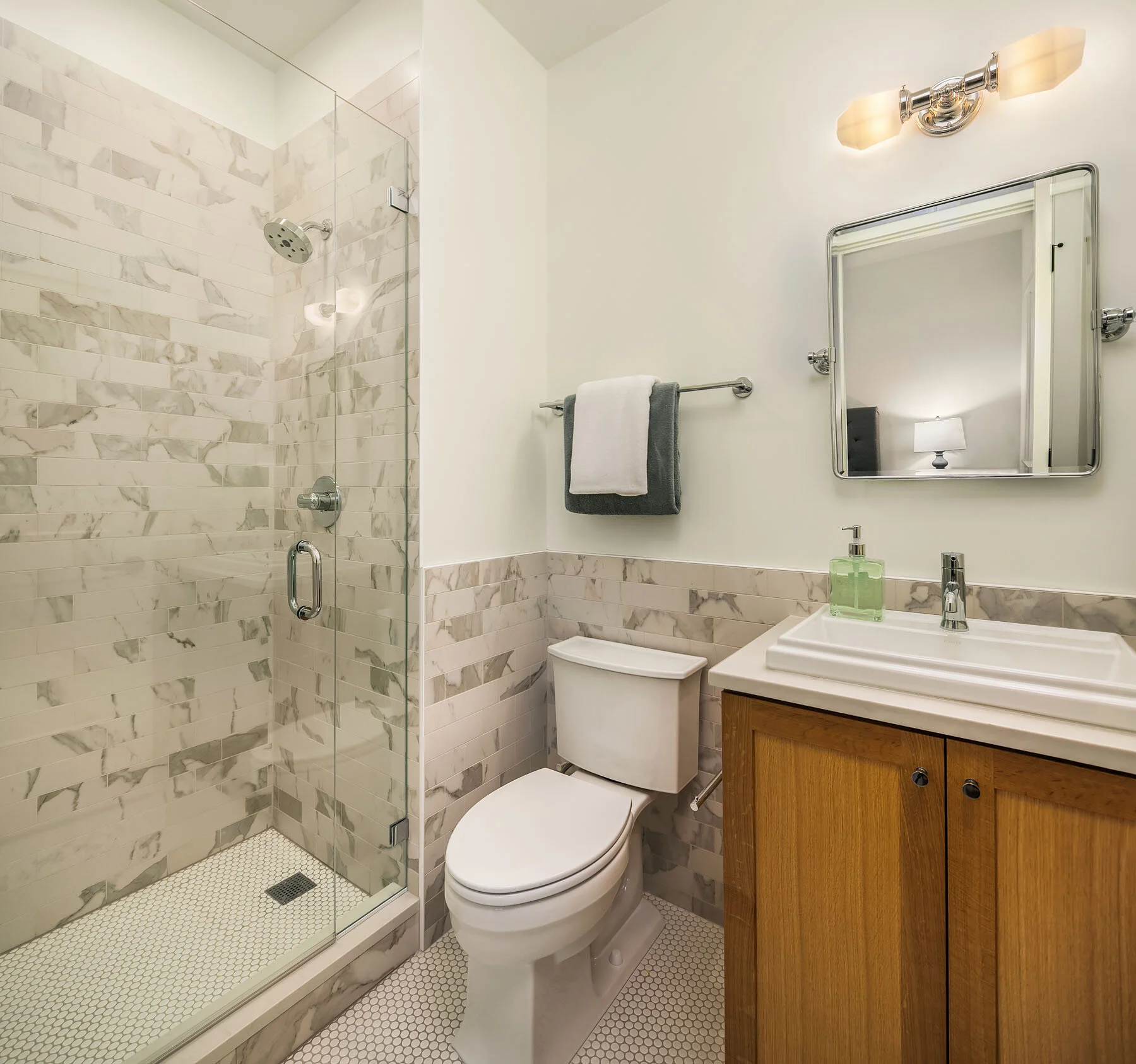 Bathroom with a glass-enclosed shower, a toilet, a wooden vanity with a sink, a rectangular mirror, a wall light fixture, and a towel rack with two towels.