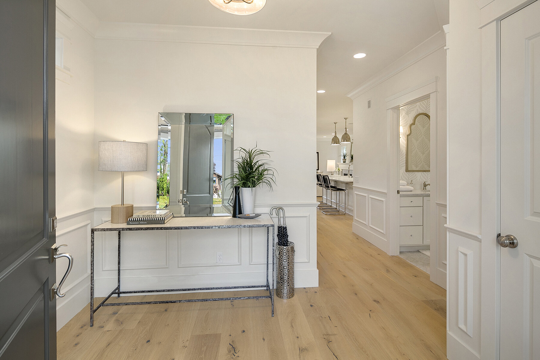 Entryway with a console table, lamp, potted plant, and mirror, leading to a kitchen and sitting area in a modern home.