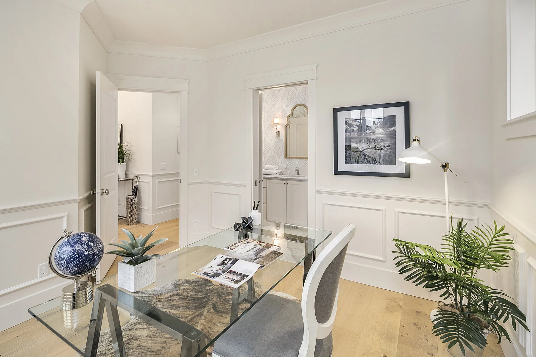A bright, modern home office with white walls, wooden flooring, a glass desk, a white and gray chair, a globe, a potted plant, and framed artwork on the wall. There is a doorway leading to a bathroom with a vanity and mirror.