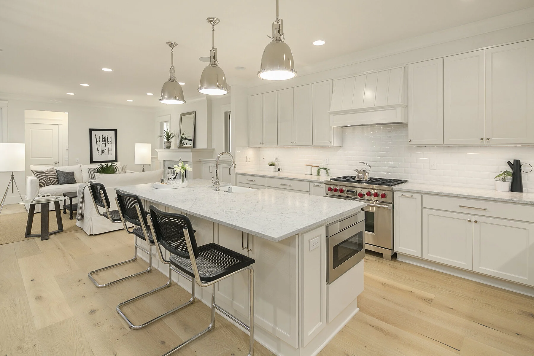 Modern white kitchen with an island, stainless steel oven, and pendant lighting, open to a living area with a sofa and lamps.