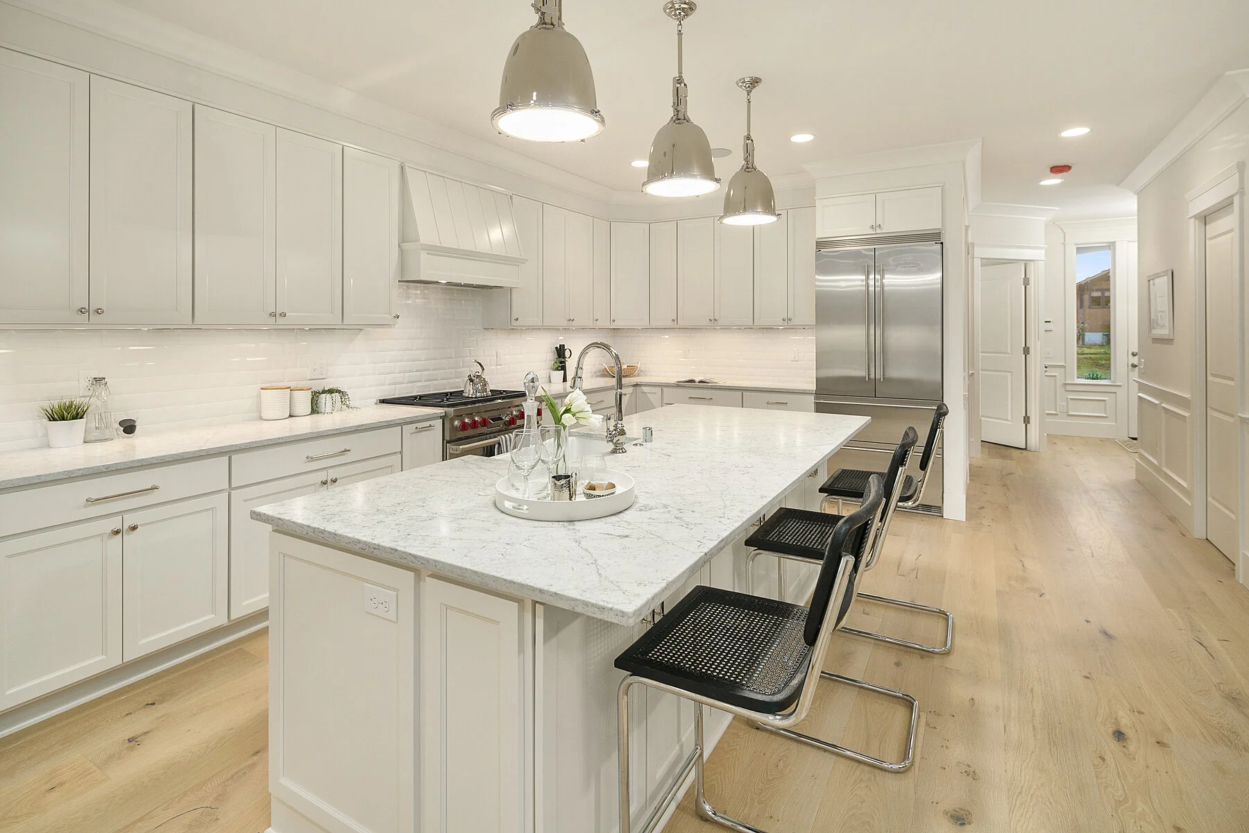 Modern kitchen with white cabinets, marble island, stainless steel appliances, light wood flooring, pendant lighting, and a view of the entry door with a window.