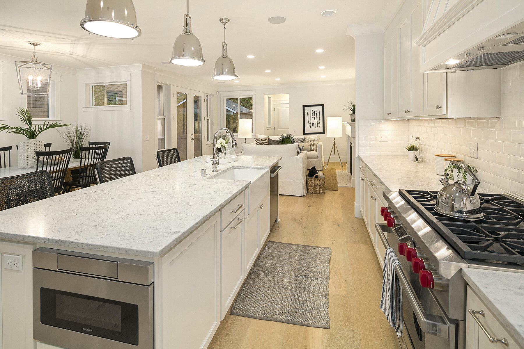 Bright, modern kitchen with white cabinets, marble countertops, stainless steel oven, and a large central island with a sink. The kitchen opens to a dining area with a dark wood table and black chairs, and further back, a living room with a white sof