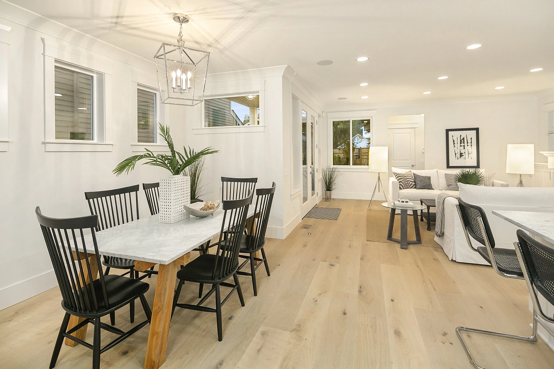 A bright, modern living and dining area with light wood flooring, white walls, and large windows. The dining area features a white marble table with black chairs, while the living area has a white sofa, assorted cushions, and side lamps. Decorative p