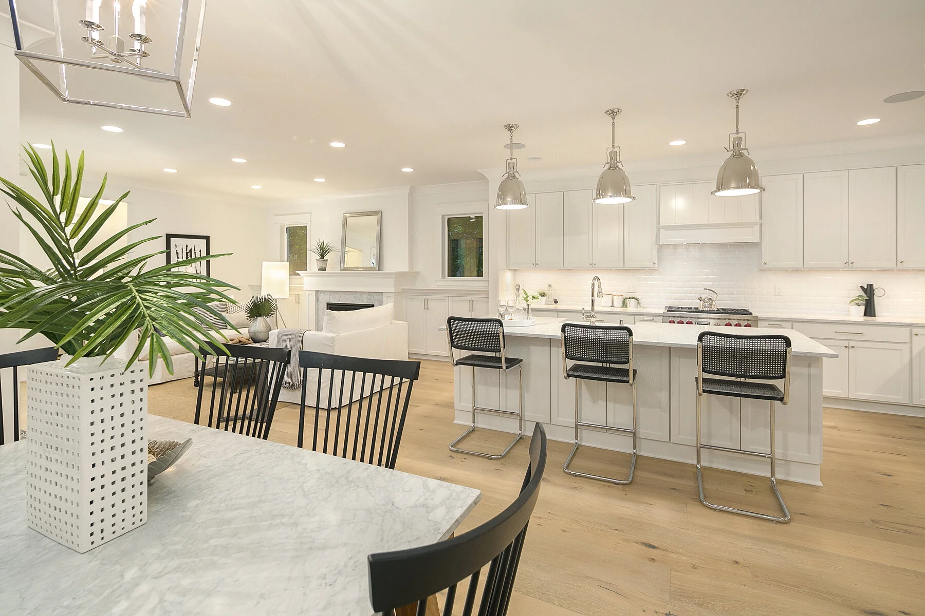 Bright, modern kitchen with white cabinets, a large marble island, and black bar stools. Close-up of dining table with a plant in a white vase in the foreground.