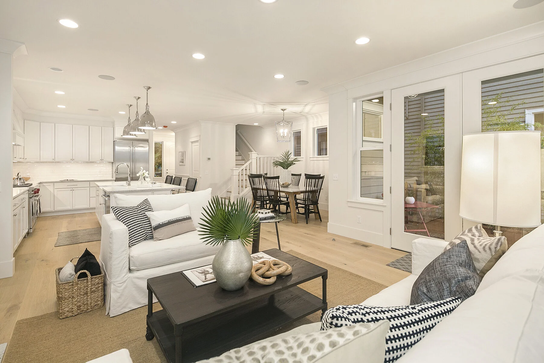 Open-concept living room and kitchen area with white walls, hardwood flooring, white cabinetry, stainless steel appliances, black dining table with six chairs, white sofas with black and patterned pillows, and a modern decor style.