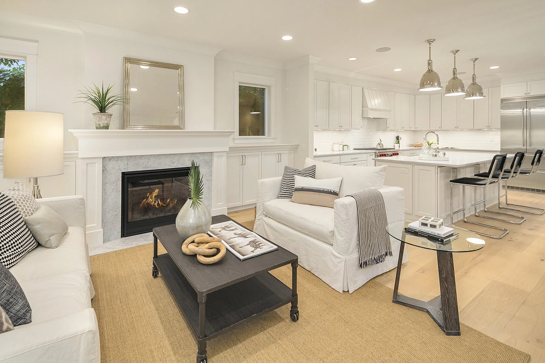 Living room with white sofas, a black coffee table, a fireplace, a glass side table, and a kitchen with a large island and bar stools.
