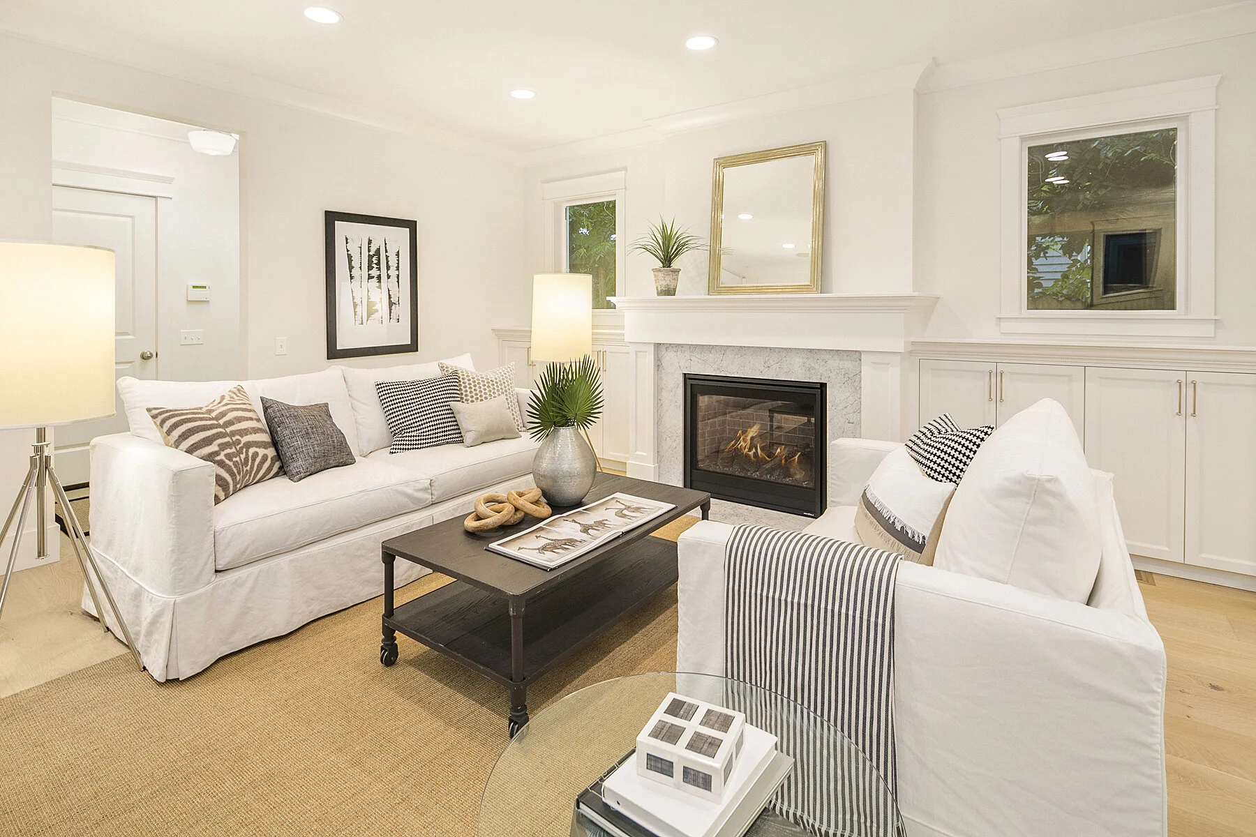 Living room with white sofas, black coffee table, fireplace, framed artwork, green plant, lamps, and windows.