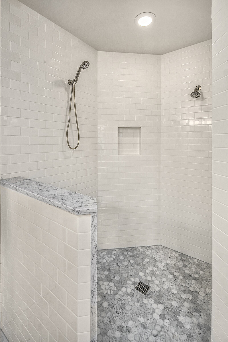 A modern walk-in shower with white subway tiles on the walls, hexagonal gray tile flooring, and two shower heads. A recessed shelf is built into the wall, and a marble half wall separates the shower area from the rest of the bathroom. A ceiling light