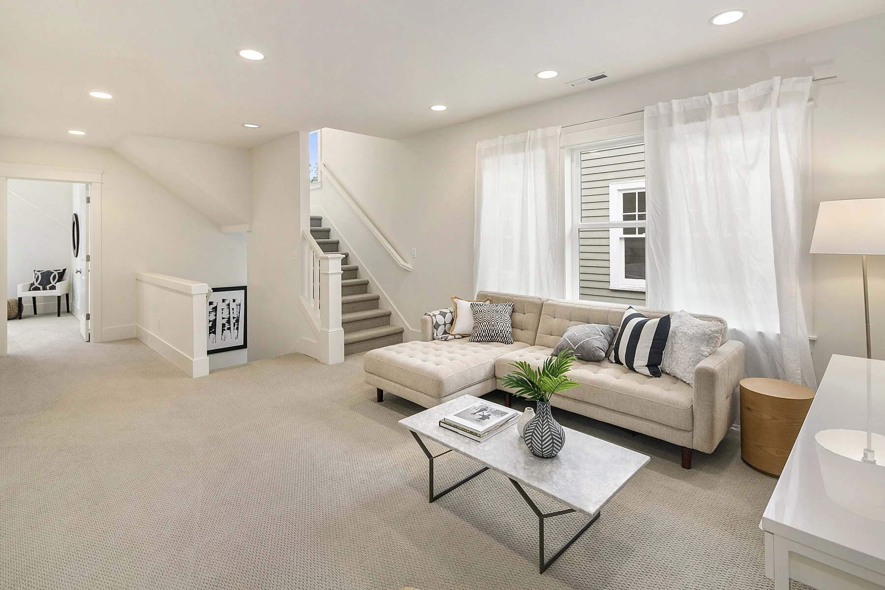 Living room with beige sectional sofa, white curtains, windows, marble coffee table with books and plant, beige carpet, and white walls.