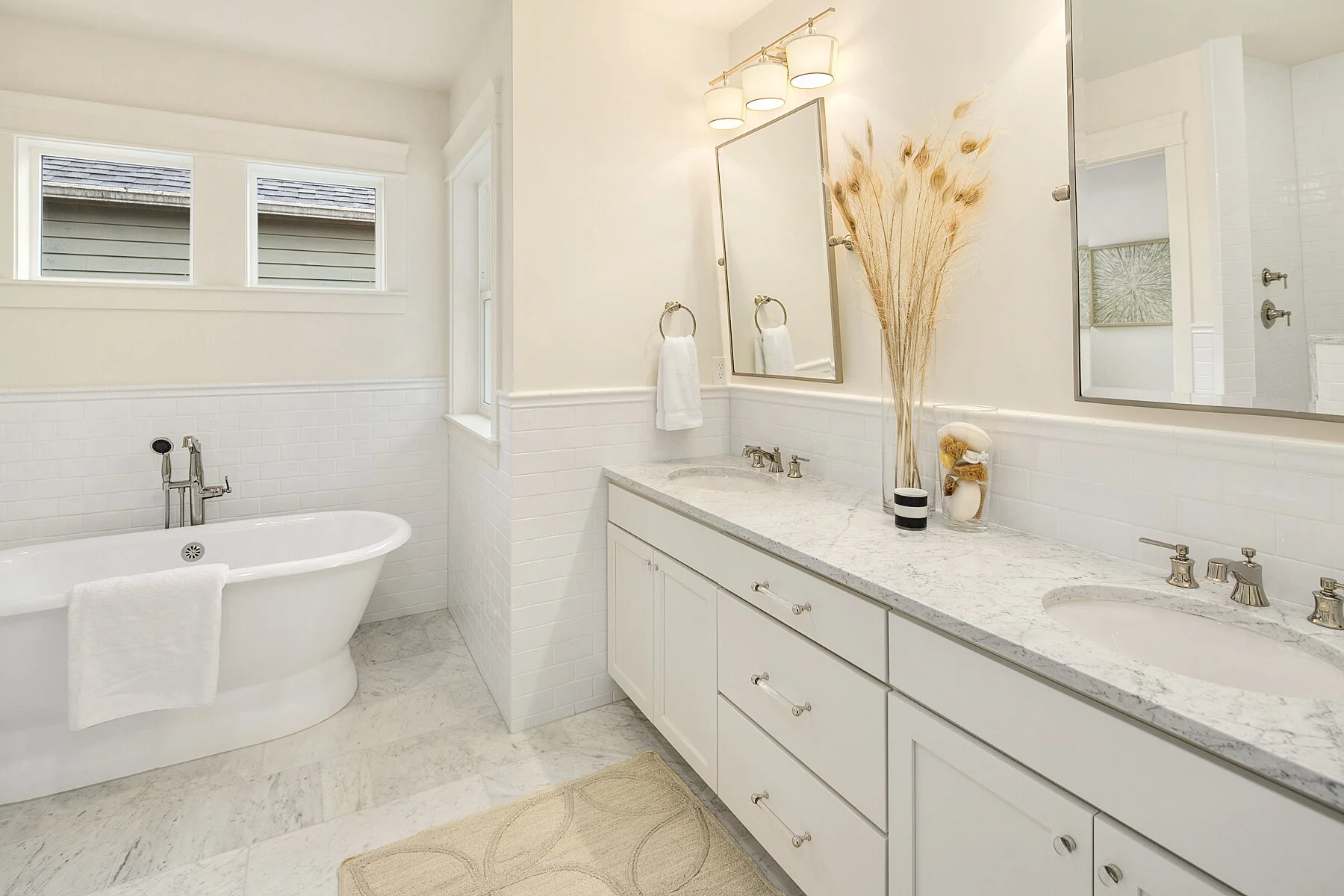 Bright bathroom with white tiled walls, a bathtub with a white towel, a double vanity with a marble countertop, two mirrors, and decorative vases and dried plants.