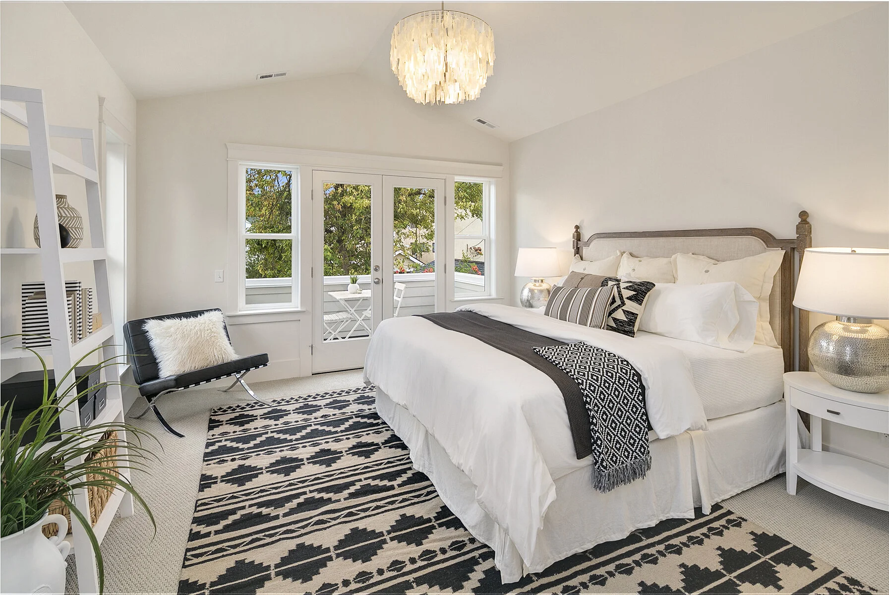 A bedroom with a large bed, white linens, and patterned pillows. There are two nightstands with lamps, a shelving unit, a black lounge chair with a fur pillow, and glass doors leading to a small balcony with outdoor furniture. The room features white