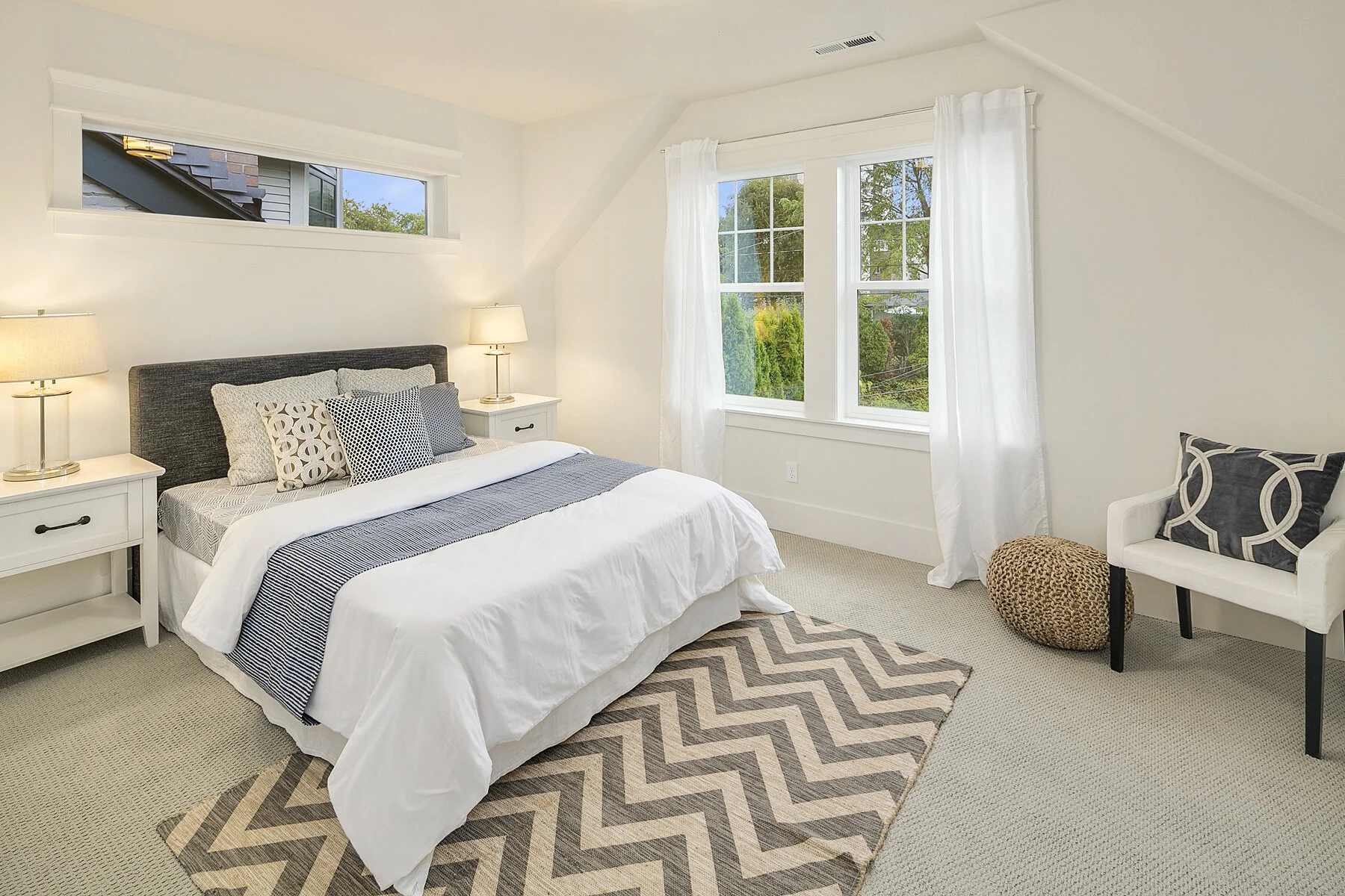 A bright bedroom with white walls, two nightstands with lamps beside a bed with a dark gray headboard, and a chevron-patterned rug on beige carpet. Large windows with white curtains reveal greenery outside, and a white chair with a black and white pi