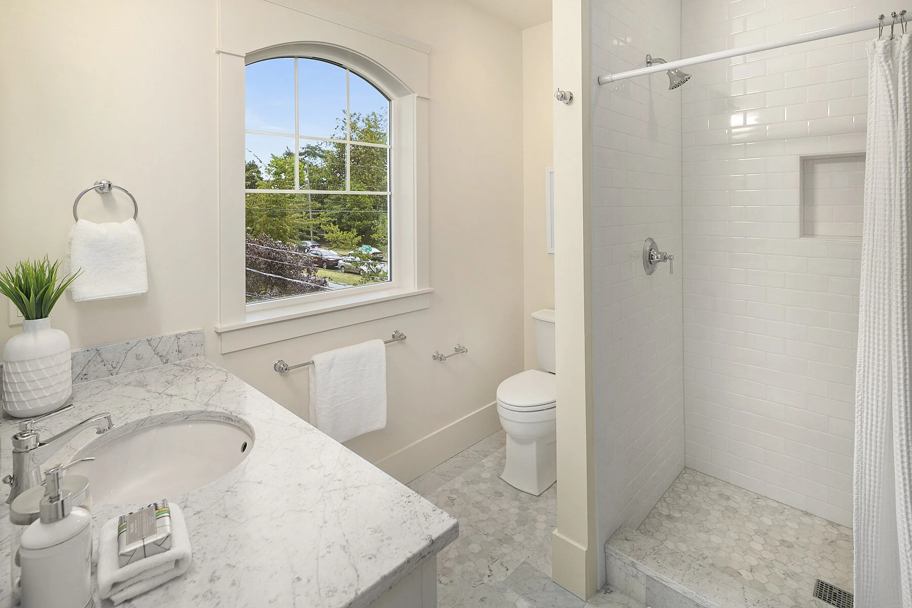 A bright bathroom with a white marble vanity, a window showing greenery outside, a toilet, and a shower with white tiles and a built-in shelf.
