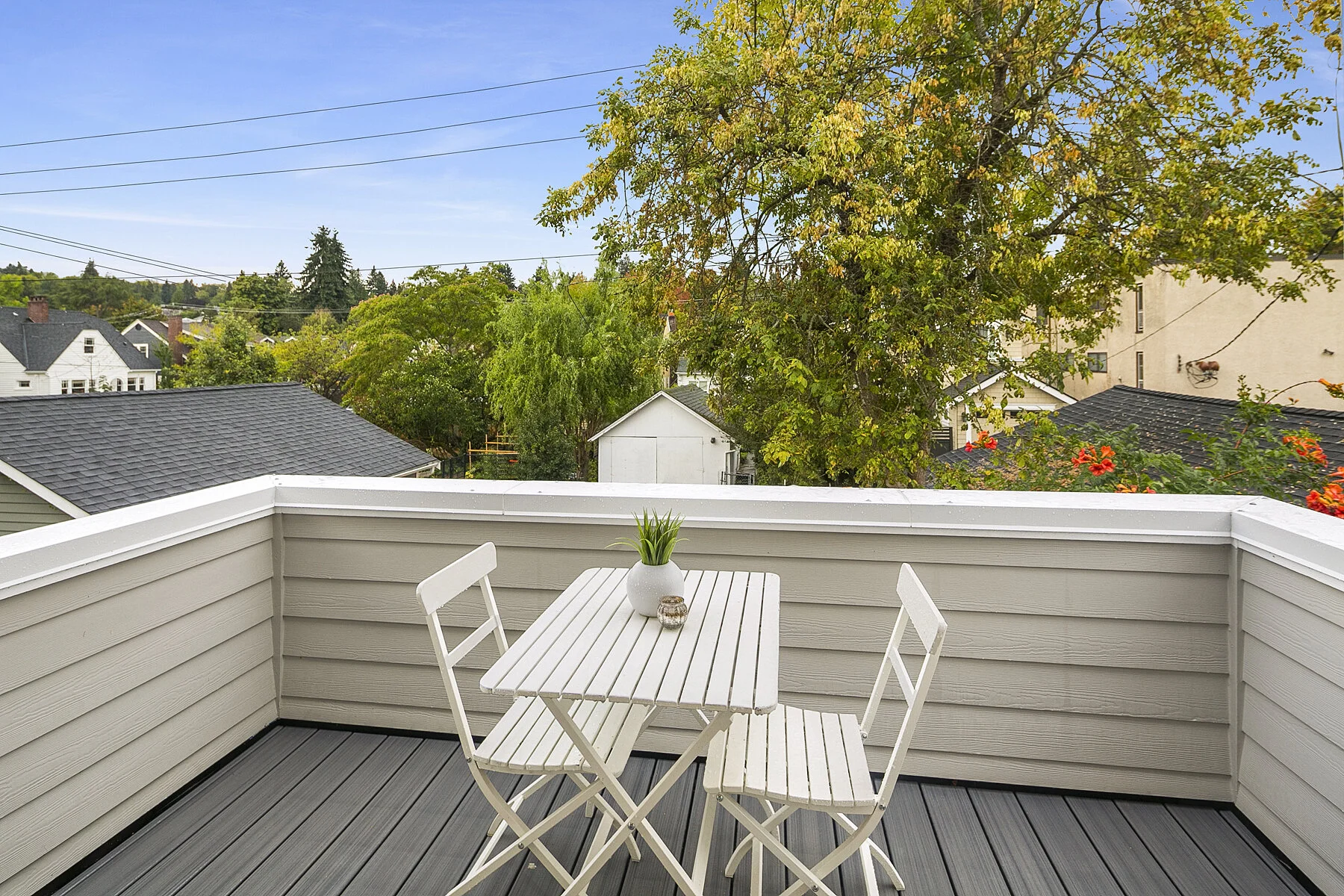Empty white outdoor patio table with two matching chairs on a wooden deck, overlooking a suburban neighborhood with trees, houses, and a clear blue sky.