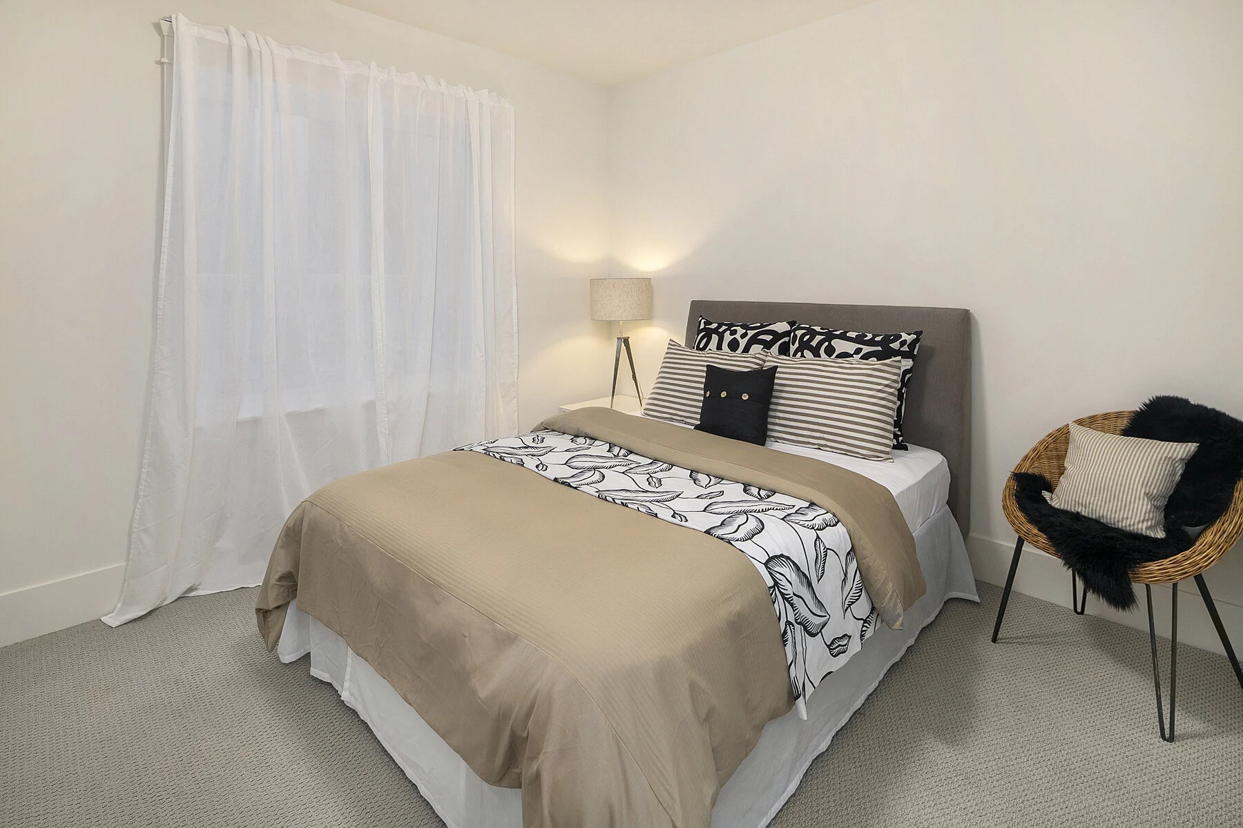 A neatly made bed with a gray headboard, beige and black pillows, a black and white patterned blanket, and a beige and white bedspread in a minimalistic bedroom. A floor lamp with a beige shade is beside the bed, and a wicker chair with a cushion and