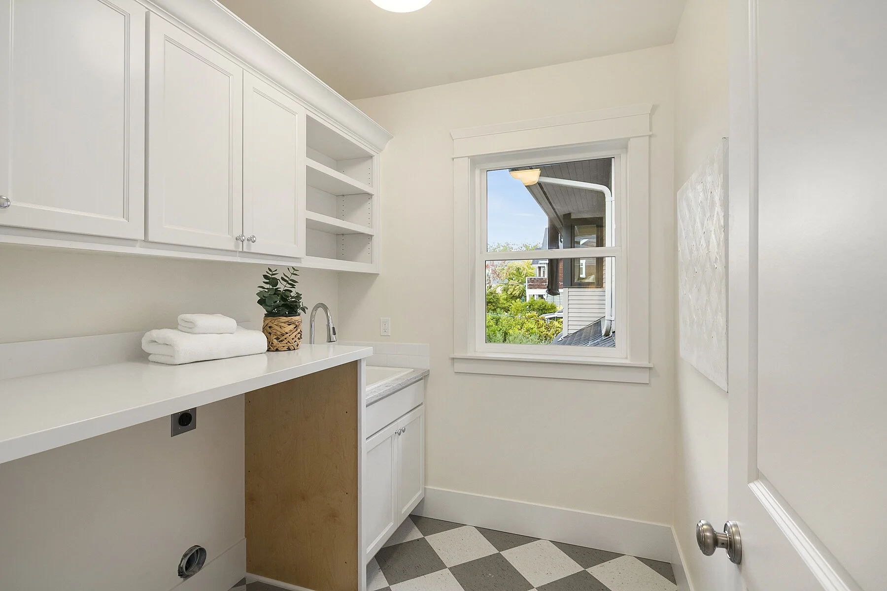 A laundry room with white cabinets, a countertop, a small sink, a window showing a view of trees and neighboring houses, and a checkered floor.