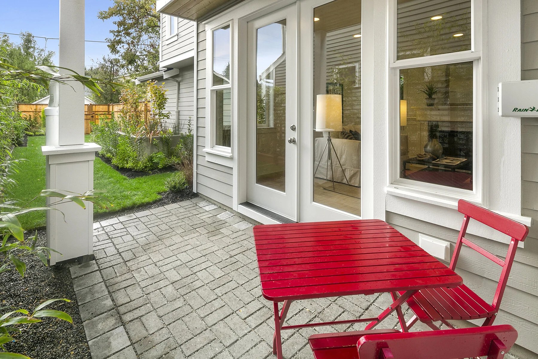 A small outdoor patio with gray brick flooring, a red metal table and two matching chairs, white siding on the house, a glass door, and window, with a well-maintained garden and green lawn in the background.