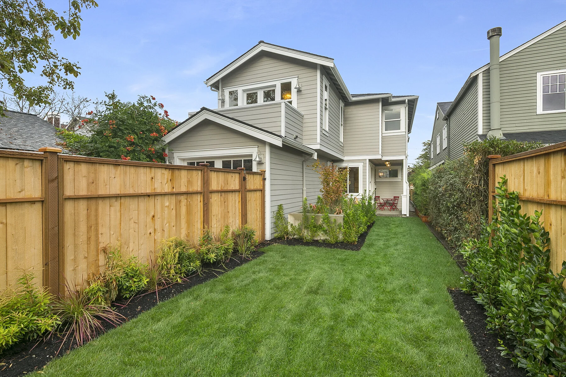Backyard with well-maintained green lawn, wooden fence on both sides, and a two-story house with gray siding and a small porch with red furniture.