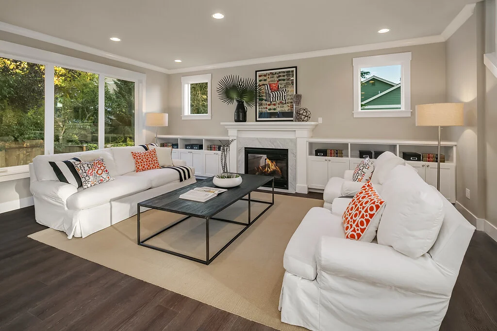 Bright living room with large windows, white sofas with orange and patterned throw pillows, fireplace, black coffee table, beige rug, and white built-in shelves.