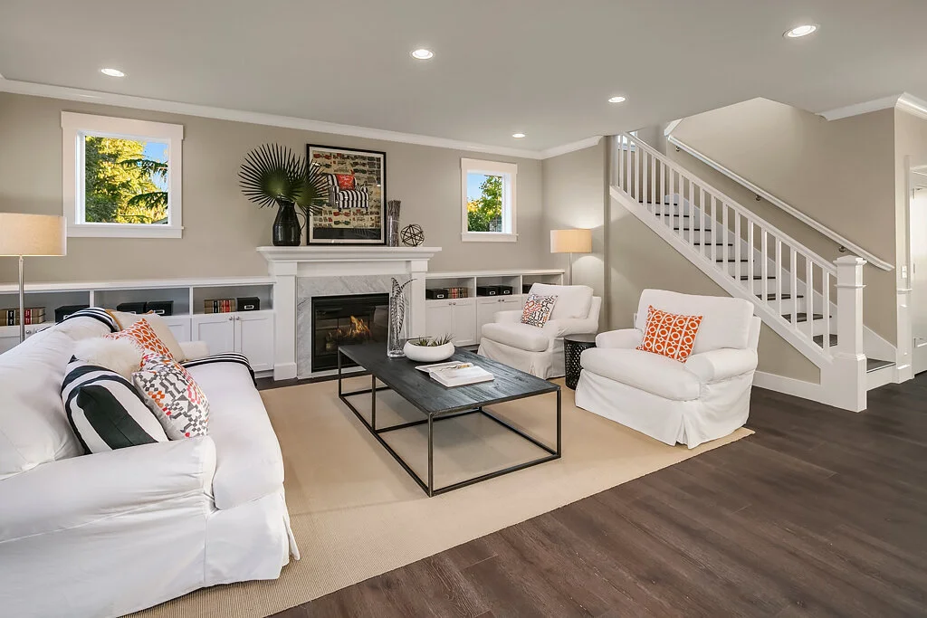 Living room with white sofa and chairs, fireplace, coffee table, built-in shelves, two small windows, and staircase with white railing.