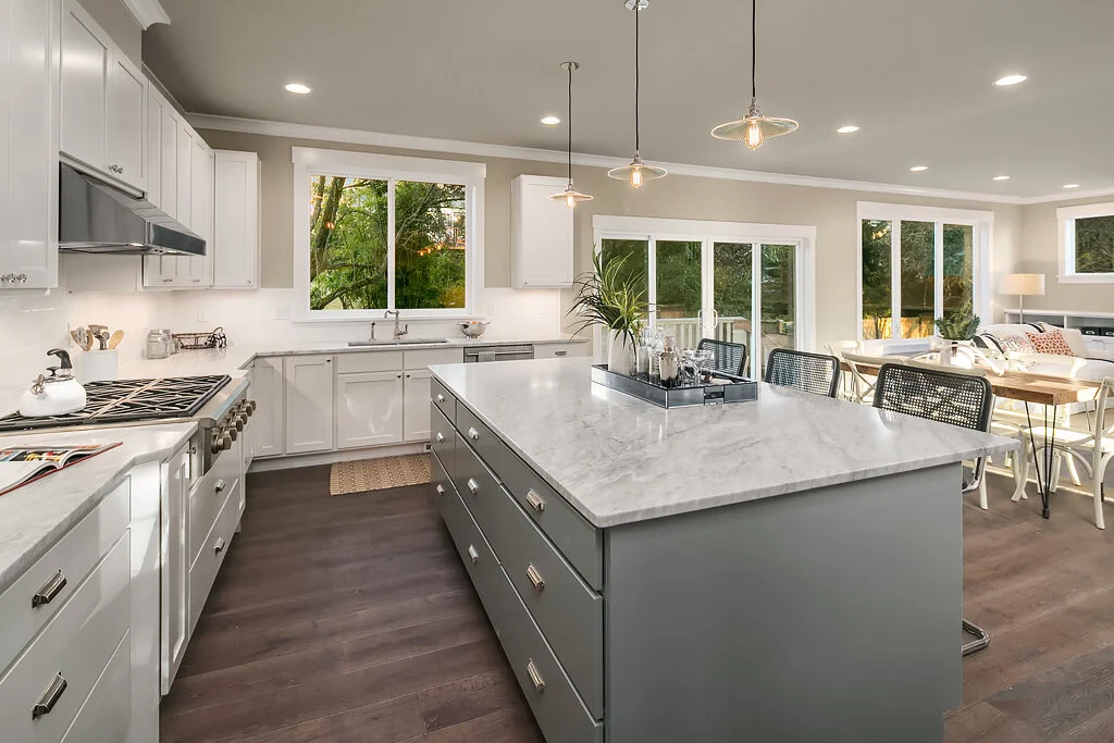 Modern kitchen with white cabinets, marble island, and large windows in a bright, open living space.