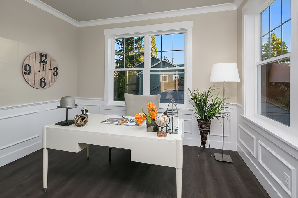 Home office with white desk, large windows, potted plant, table lamp, and wall clock.