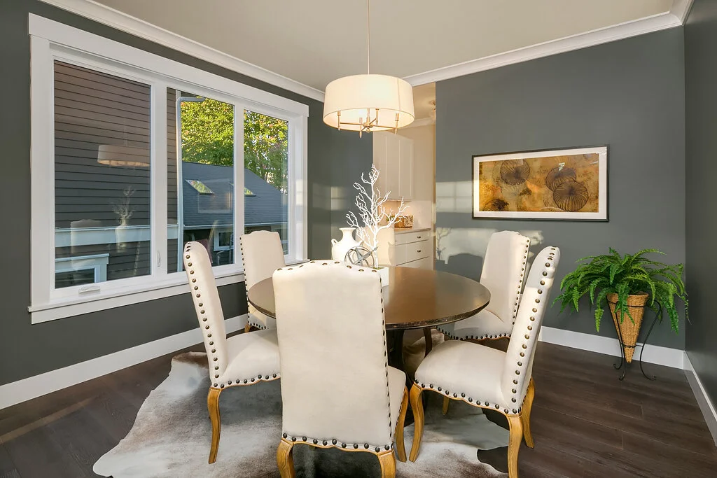 Dining room with a round wooden table, six white upholstered chairs with nailhead trim, a large window, a framed artwork on a green wall, a potted fern, and a hanging light fixture.