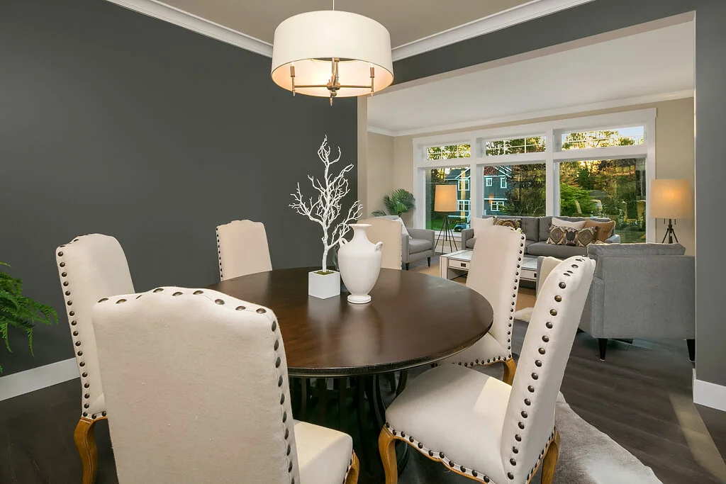A dining room with a round wooden table surrounded by six beige upholstered chairs with nailhead trim. The table has white vases and a decorative branch centerpiece. The room opens to a living area with sofas and large windows showing a wooded outdoo