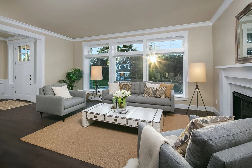 Bright living room with a large window, gray sofas, a coffee table, and lamps, with a view of a house and trees outside.