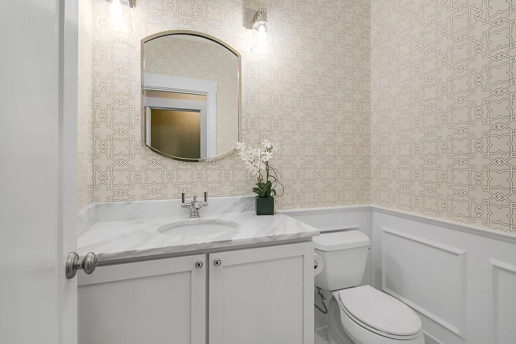 A clean bathroom with a marble countertop sink, a decorative mirror, a white toilet, and a wallpaper with a geometric pattern, accented with a small potted orchid.