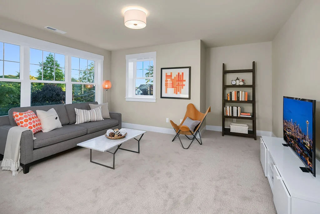 Living room with a gray sofa decorated with white and patterned pillows, a white marble coffee table with decorative objects, an orange butterfly chair, a black ladder-style bookshelf with books and decorations, a flat-screen TV on a white media cons